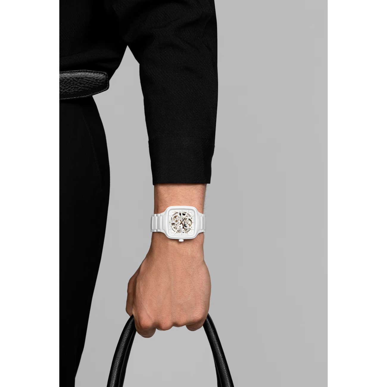 White square skeleton wristwatch with ceramic bracelet on wrist, holding a black bag handle